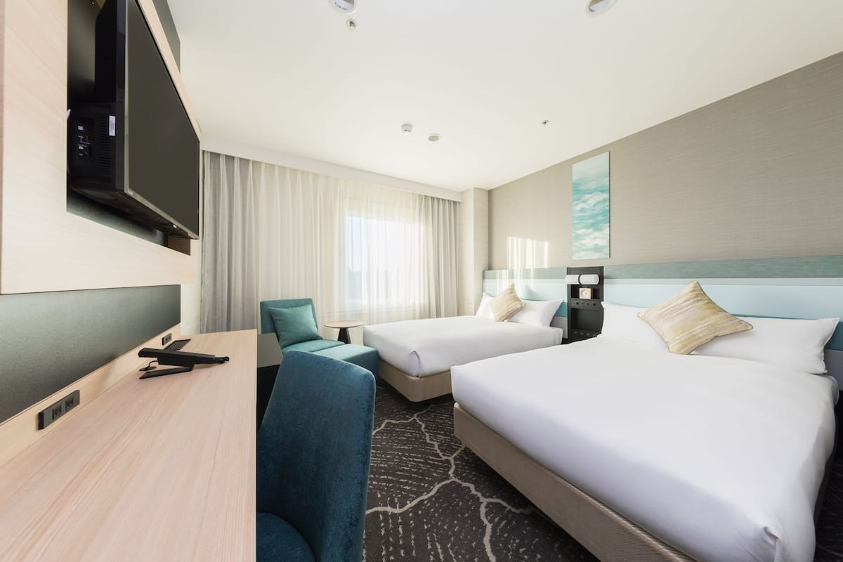 Ein Hotelzimmer mit zwei Betten, einem Schreibtisch, einem Stuhl, einem Fernseher und einem Fenster mit Vorhängen.