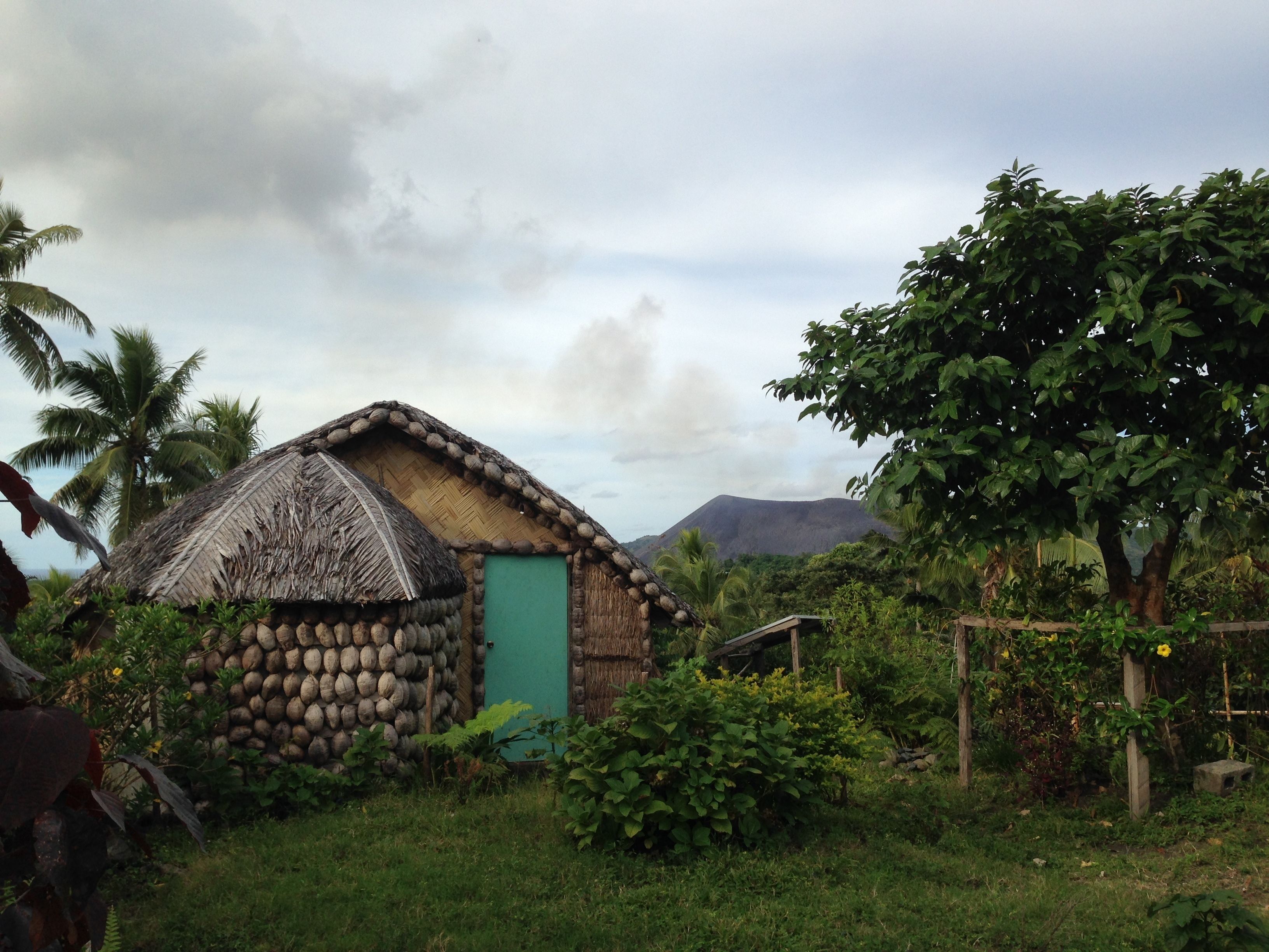 A rustic hut with a thatched roof, surrounded by lush greenery and a mountain in the background.