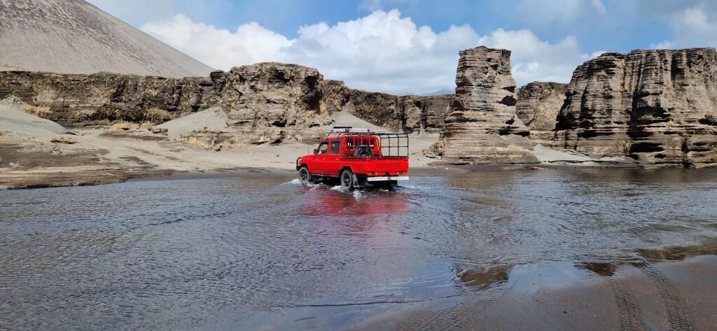 Una camioneta roja circulando por una extensión de agua poco profunda en terreno pedregoso.