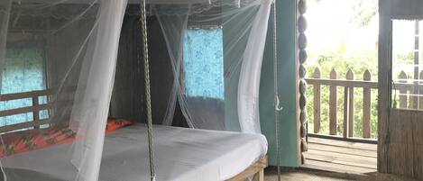 A bed with a mosquito net, a wooden floor, and a view of greenery outside.