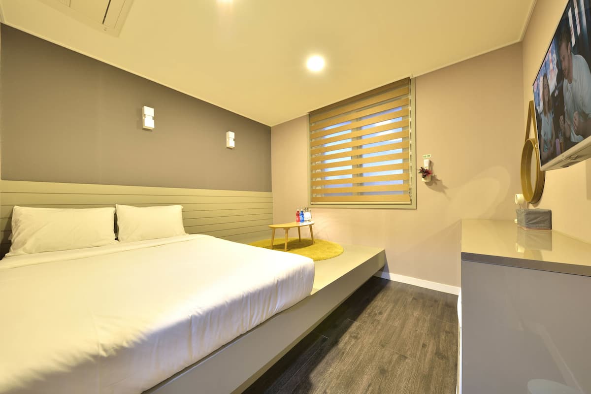 A hotel room with a bed, a small table with a yellow rug, a window with blinds, and a TV on a stand.