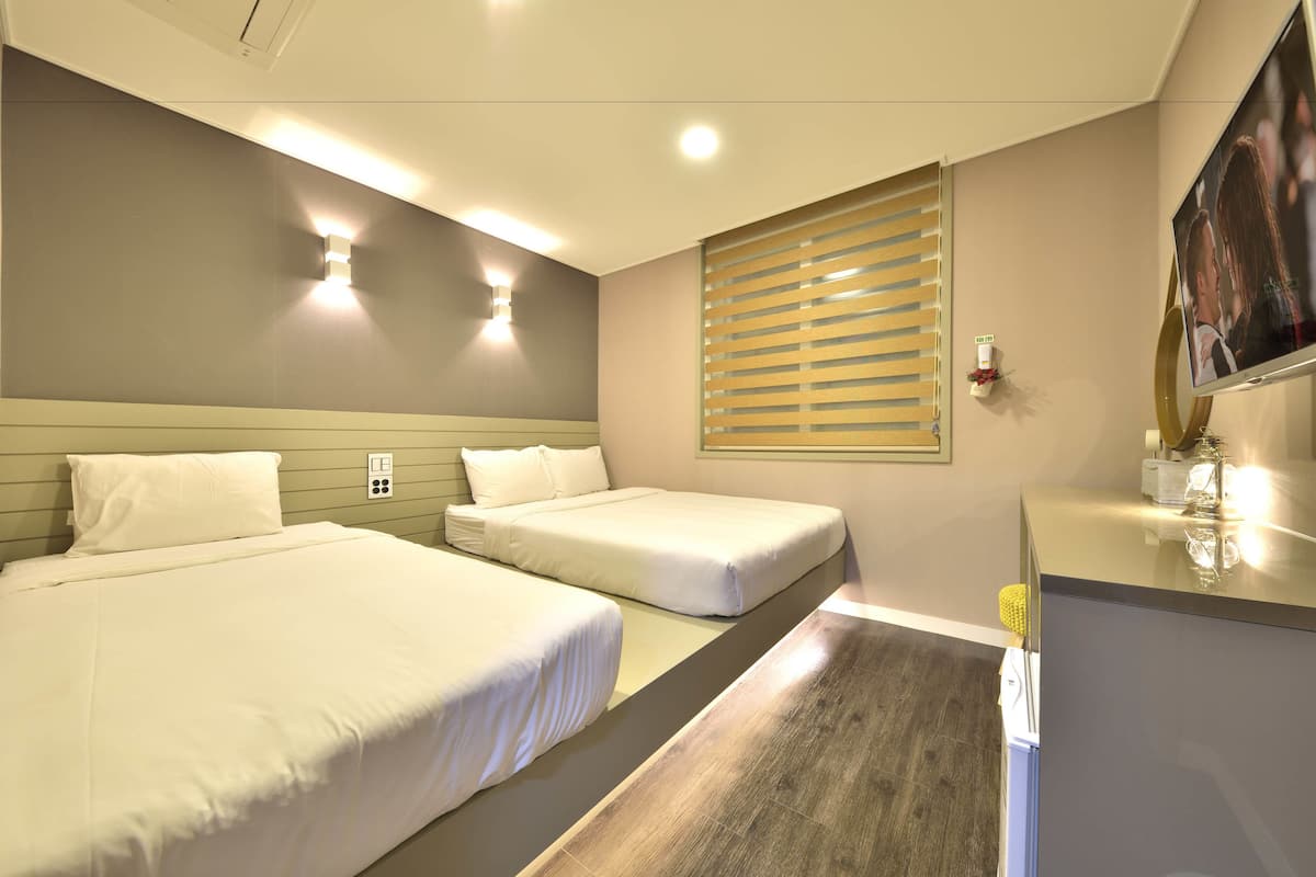 A hotel room with two beds, a small desk, a TV, and a window with blinds.