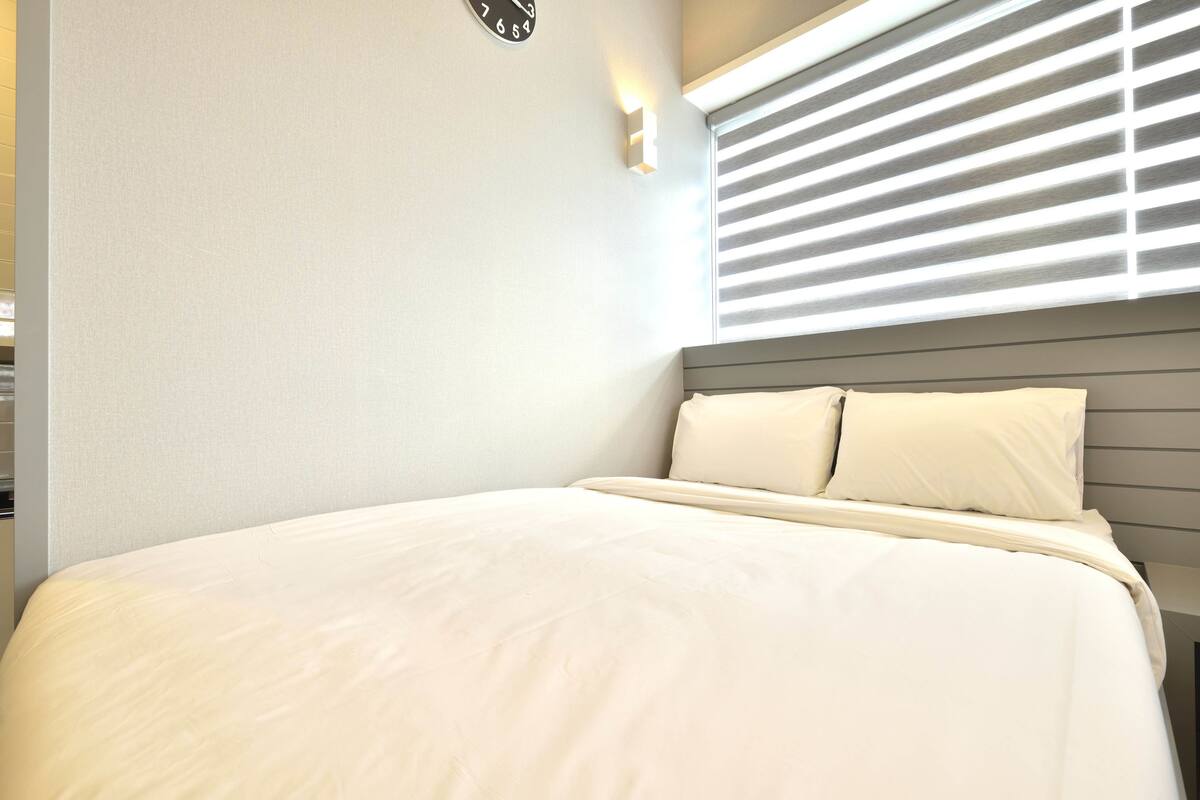 A neatly made bed with a headboard, a wall clock, and a window with blinds.