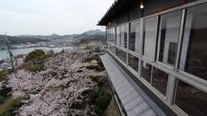 A traditional building with large windows overlooking a river and cherry blossoms. - Onomichi Guest House Miharashi-tei - Hostel (Onomichi)