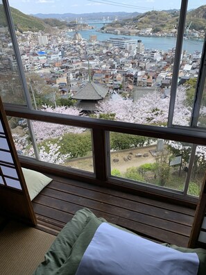 A room with a large window overlooking a cityscape with traditional and modern buildings, a river, and cherry blossoms. - Onomichi Guest House Miharashi-tei - Hostel (Onomichi)