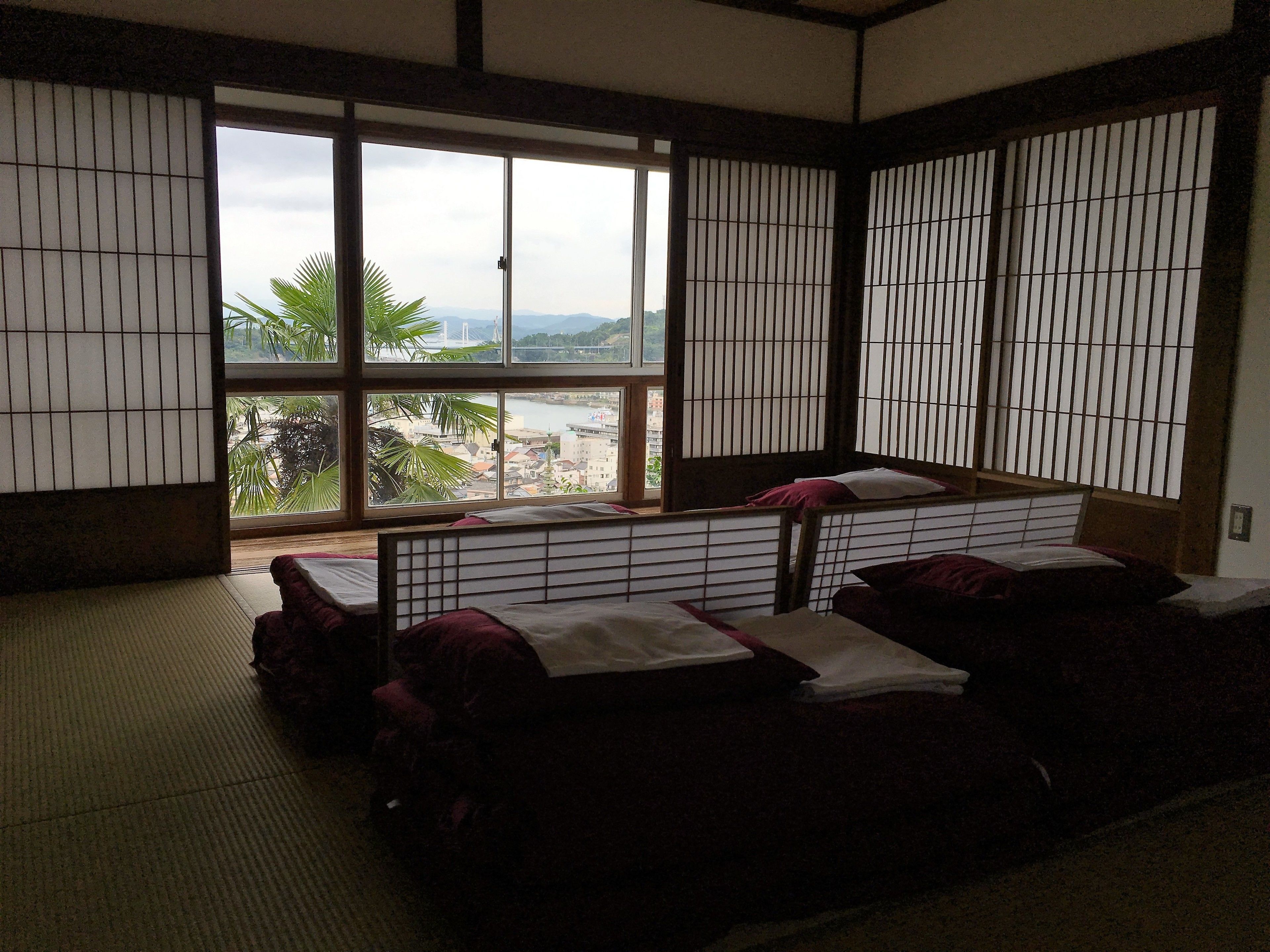 A traditional Japanese room with tatami flooring, a low bed with pillows, and a large window offering a view of a river and distant buildings.