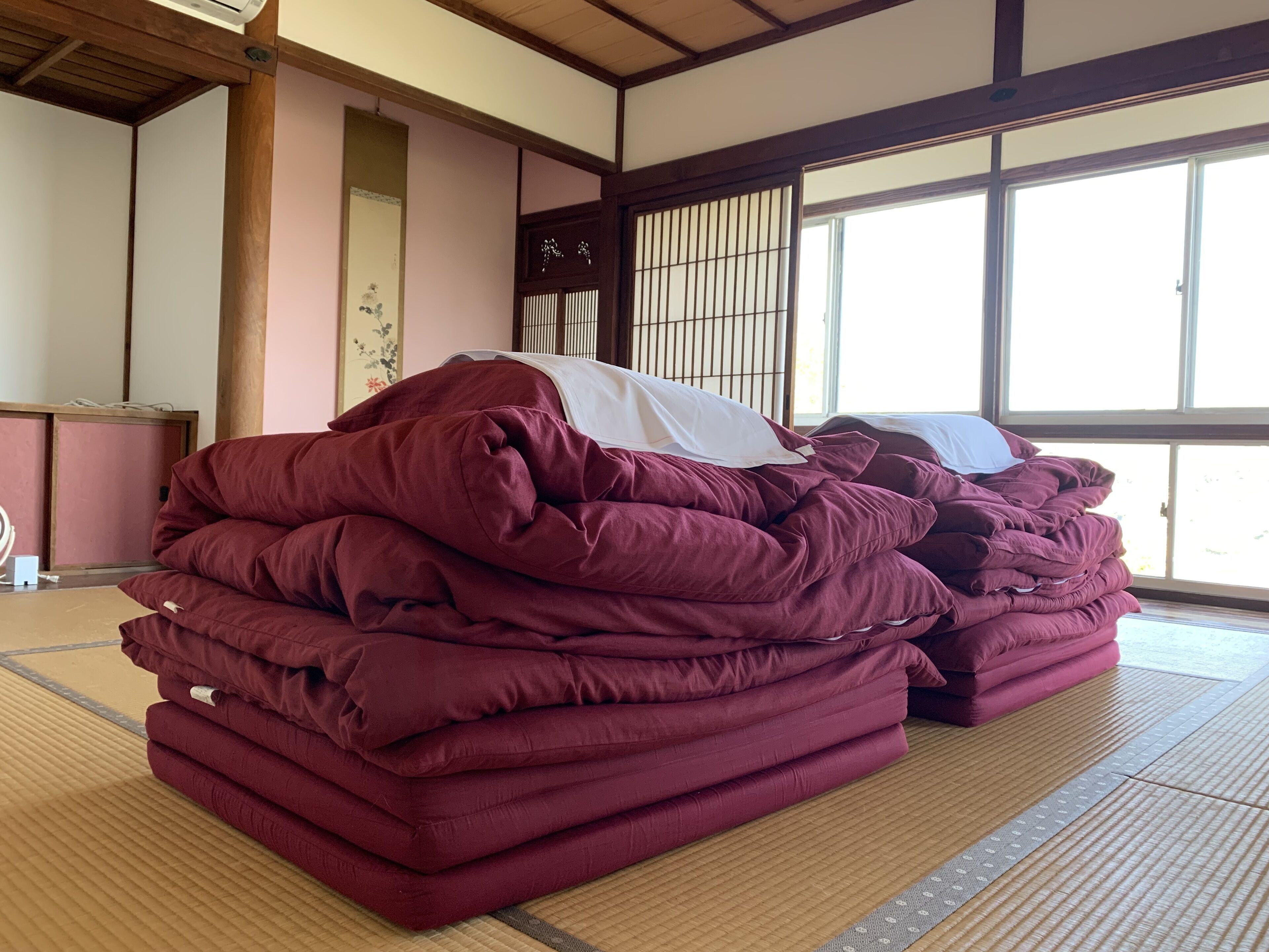 A room with tatami flooring, a large window, and a stack of red bedding.