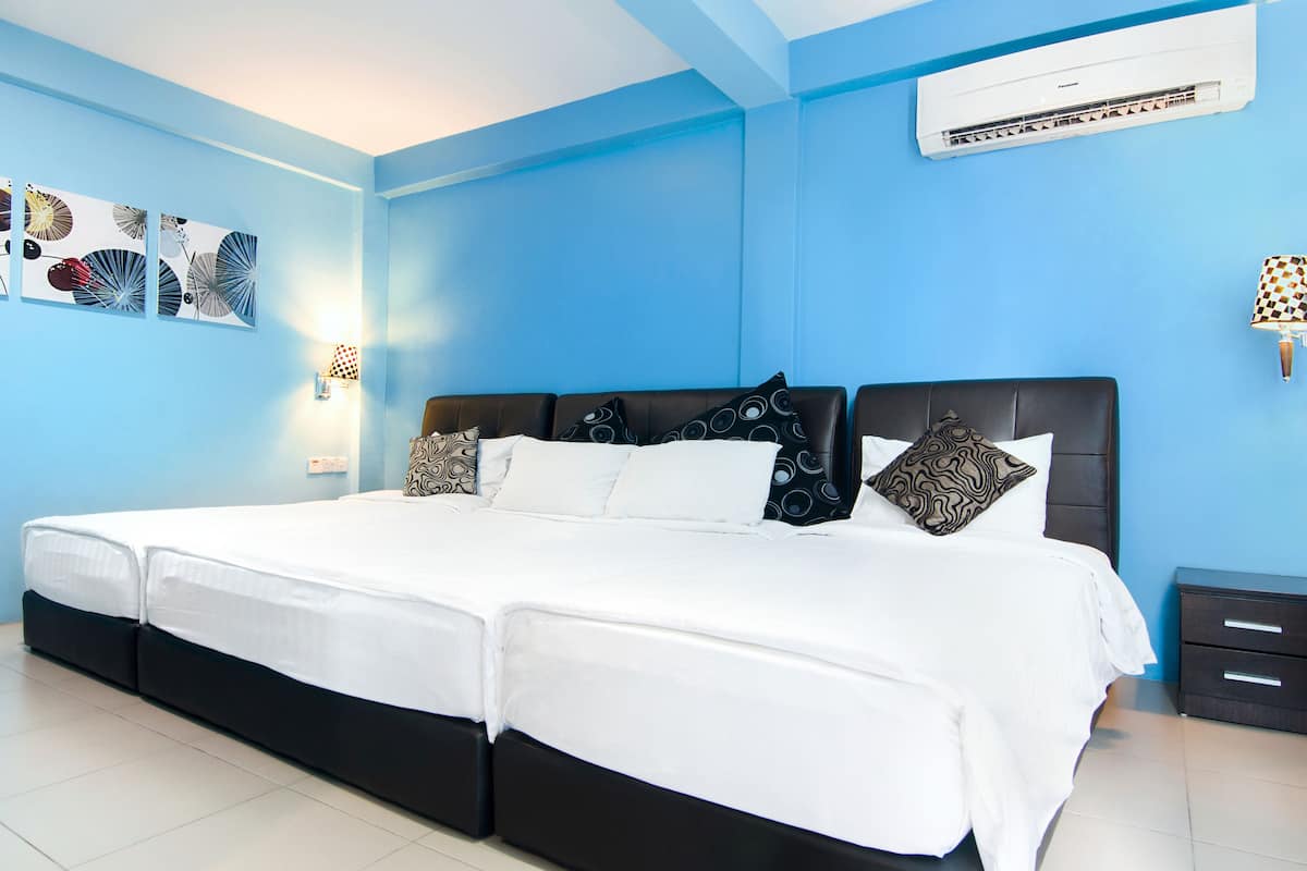 A large bed with a black headboard, white bedding, and patterned pillows. There is a wall-mounted air conditioner and a small bedside table with drawers.