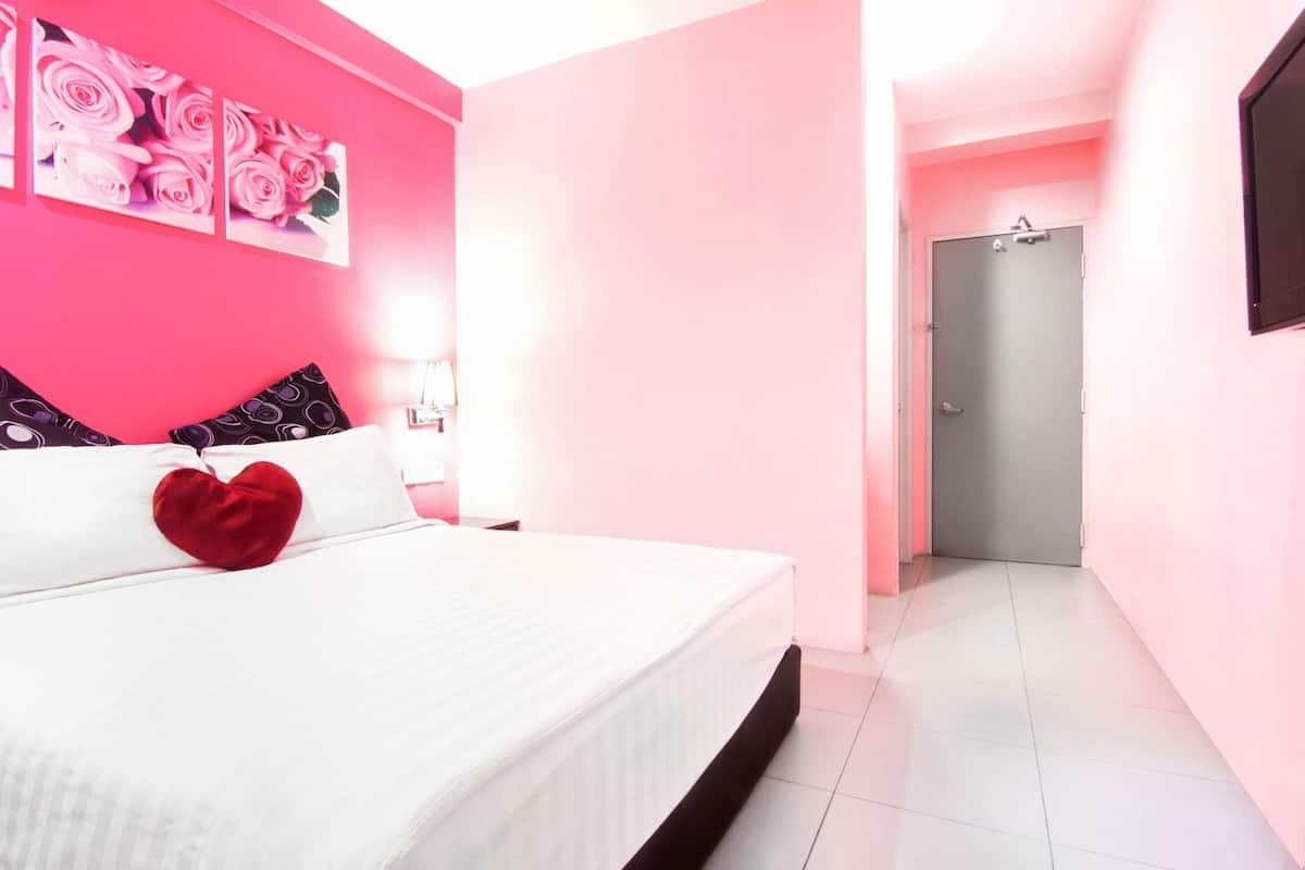 A hotel room with a bed, a heart-shaped pillow, a pink wall, and a door leading to another room.