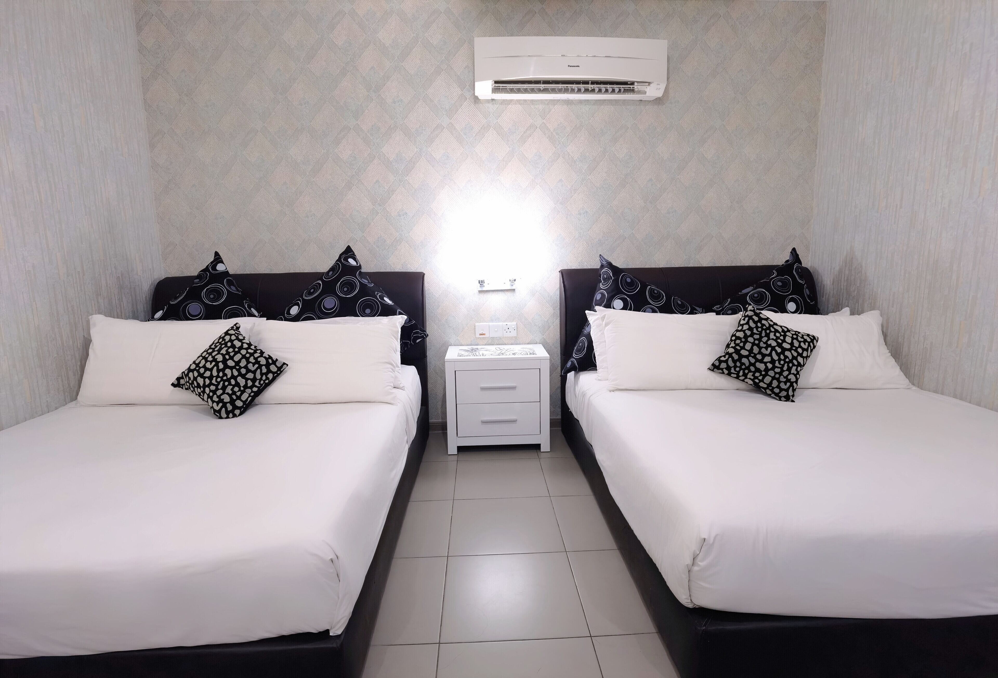 Ein Hotelzimmer mit zwei Betten, jeweils mit weißer Bettwäsche und gemusterten Kissen, einem Nachttisch mit Lampe und einer Klimaanlage an der Wand.