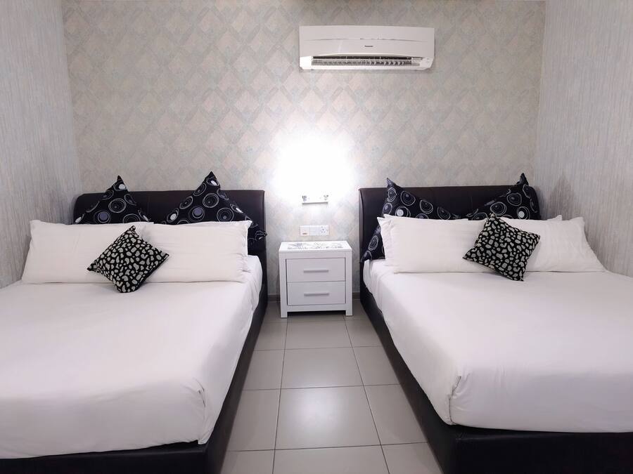 Ein Hotelzimmer mit zwei Betten, jeweils mit weißer Bettwäsche und gemusterten Kissen, einem Nachttisch mit Lampe und einer Klimaanlage an der Wand.