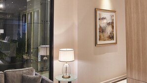 A modern living room with a sofa, a glass table with a lamp, a painting on the wall, and a ceiling light fixture. - Corsica Hotel (Kulai)