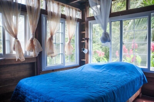 A bedroom with a large bed, sheer curtains, and a view of lush greenery outside.
