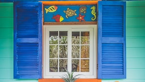 Quarto com janela exibindo arte colorida, duas cadeiras de madeira, uma mesinha com uma planta e um banquinho de madeira.