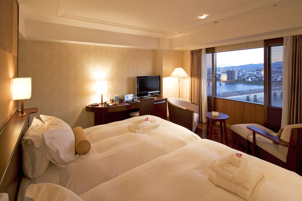 Habitación de hotel con una cama grande, un escritorio con televisor, una silla y vistas a un paisaje urbano por la ventana.