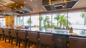 A modern restaurant with a long bar, multiple chairs, and large windows offering a view of palm trees and a cityscape. - Miyazaki Kanko Hotel (Miyazaki)