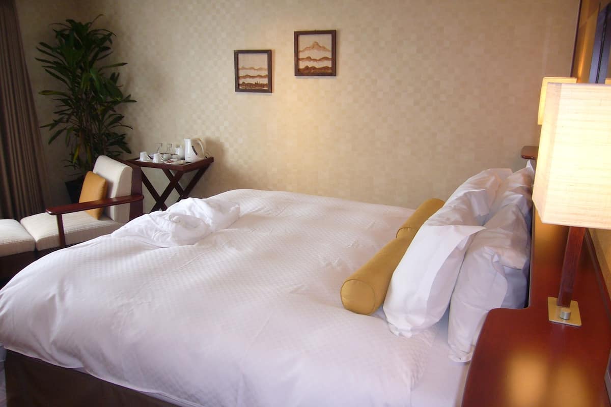 Una cama bien hecha con sábanas blancas, una mesita de noche de madera con una lámpara, una silla y una pequeña mesa con una planta y una cafetera.