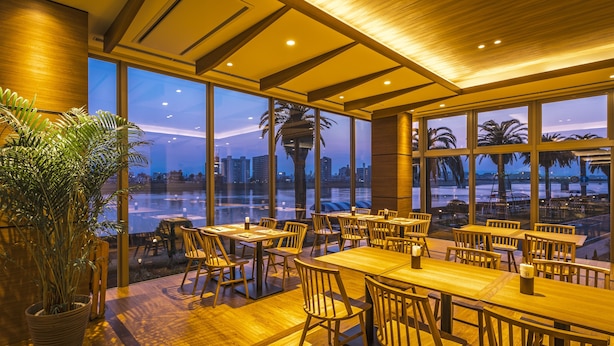 A restaurant with wooden tables and chairs, large windows with a view of the cityscape, and a potted plant. - Miyazaki Kanko Hotel (Miyazaki)