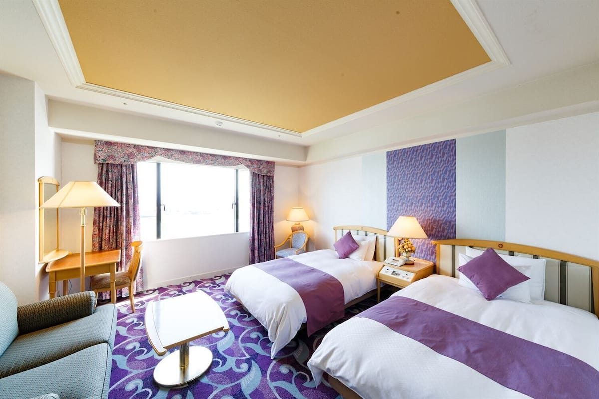 Habitación de hotel con dos camas, un escritorio, una silla y una ventana con cortinas.