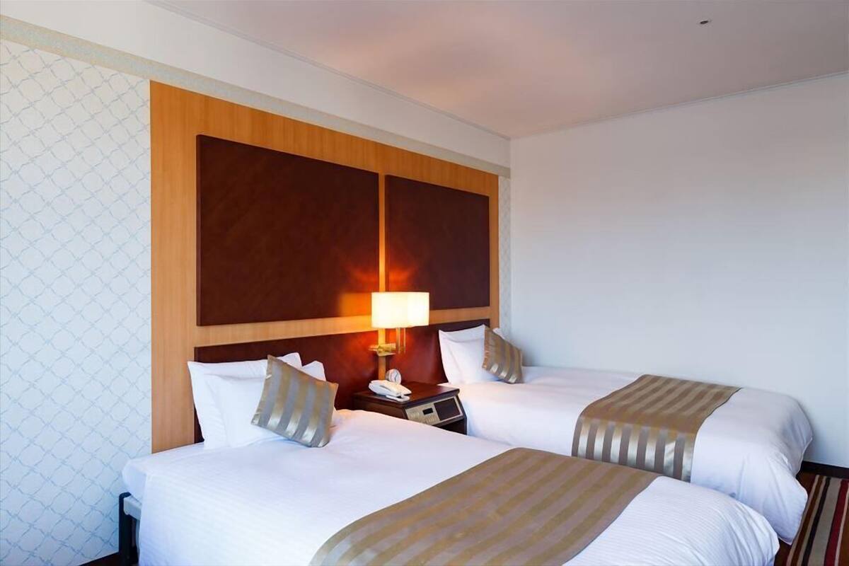Habitación de hotel con dos camas, cabecero de madera, mesita de noche con lámpara y pared con estampado.