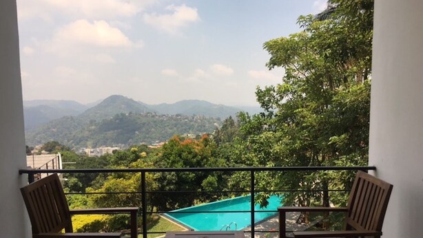 A balcony with two wooden chairs and a small table, overlooking a swimming pool and lush greenery with distant mountains. - The Kandyan Villa (Kandy)