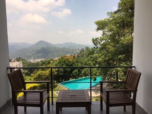 A balcony with two wooden chairs and a small table, overlooking a swimming pool and lush greenery with distant mountains. - The Kandyan Villa (Kandy)