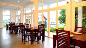 A dining area with wooden tables and chairs, large windows with a view of greenery and a building. - The Kandyan Villa (Kandy)