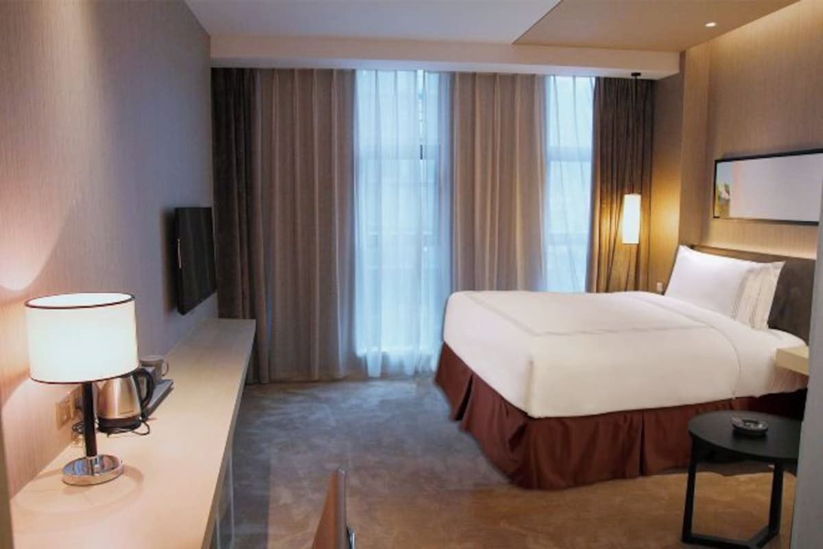 A hotel room with a bed, a desk with a lamp, a TV, and a window with curtains.