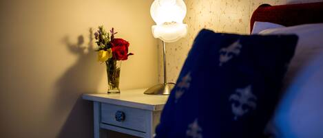 A bedroom with a bedside table, a lamp, a vase with flowers, and a bed with pillows.