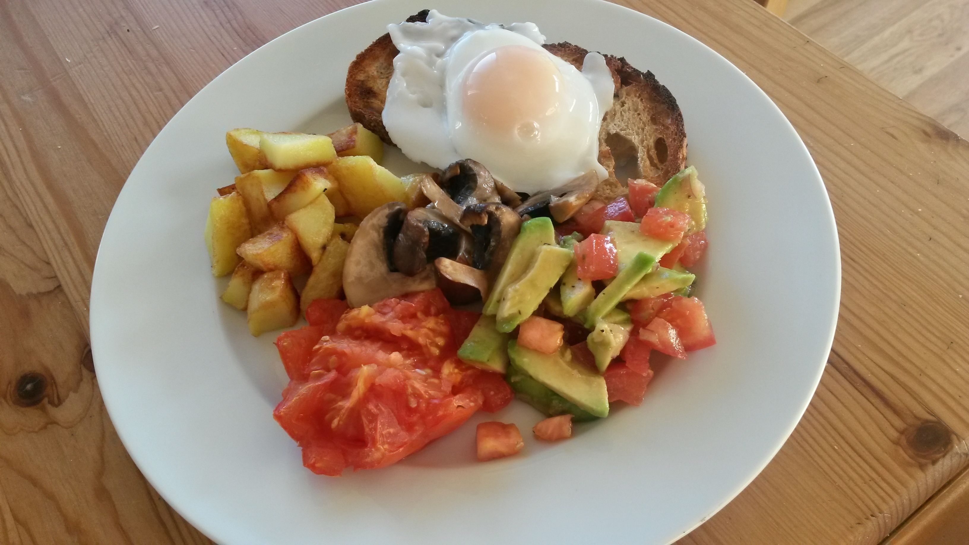 Un plato con tostadas, huevo frito, patatas en cubos, champiñones, aguacate y ensalada de tomate.