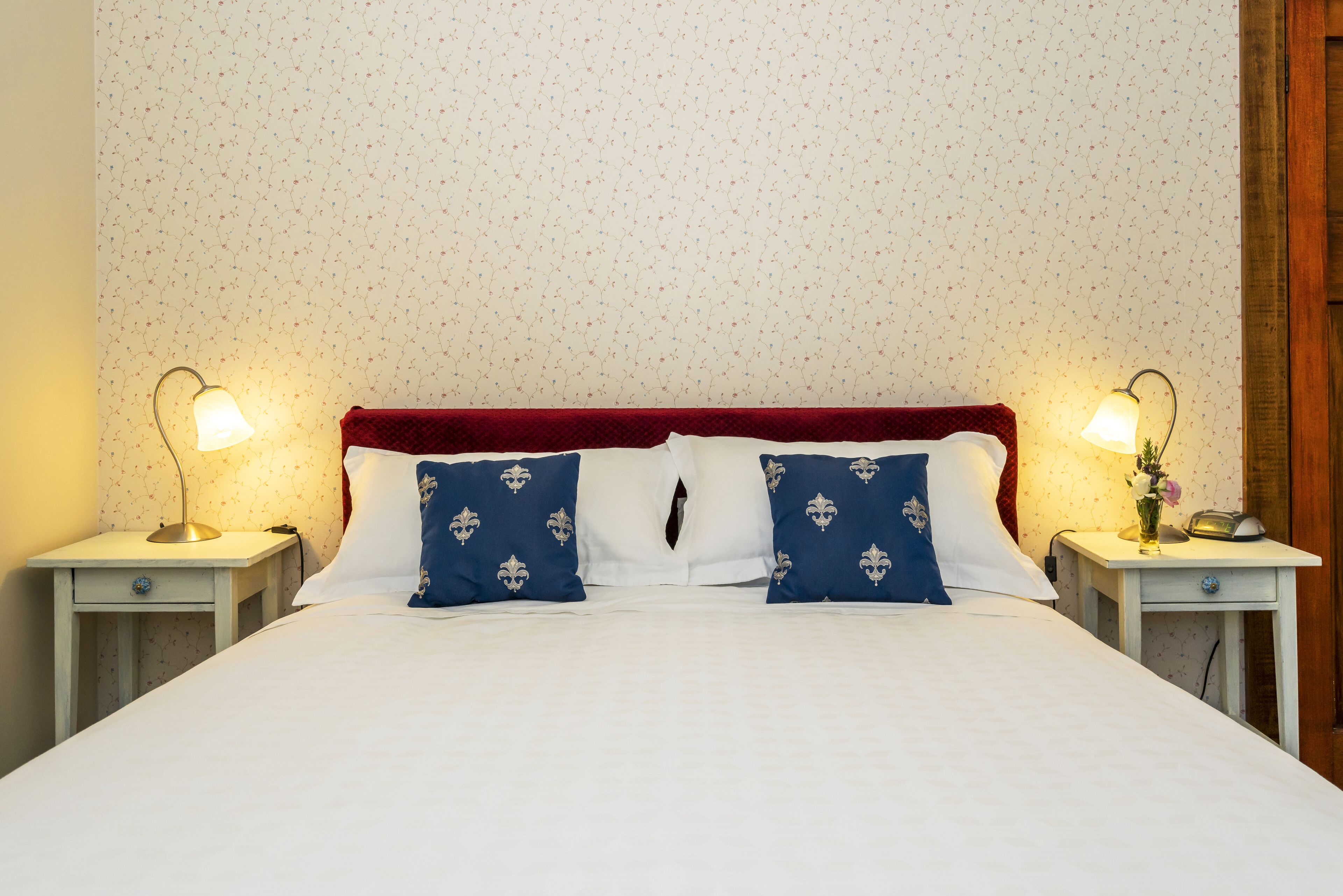 A neatly made bed with white linens and two blue pillows with fleur-de-lis patterns, flanked by two bedside tables with lamps and a small vase of flowers.