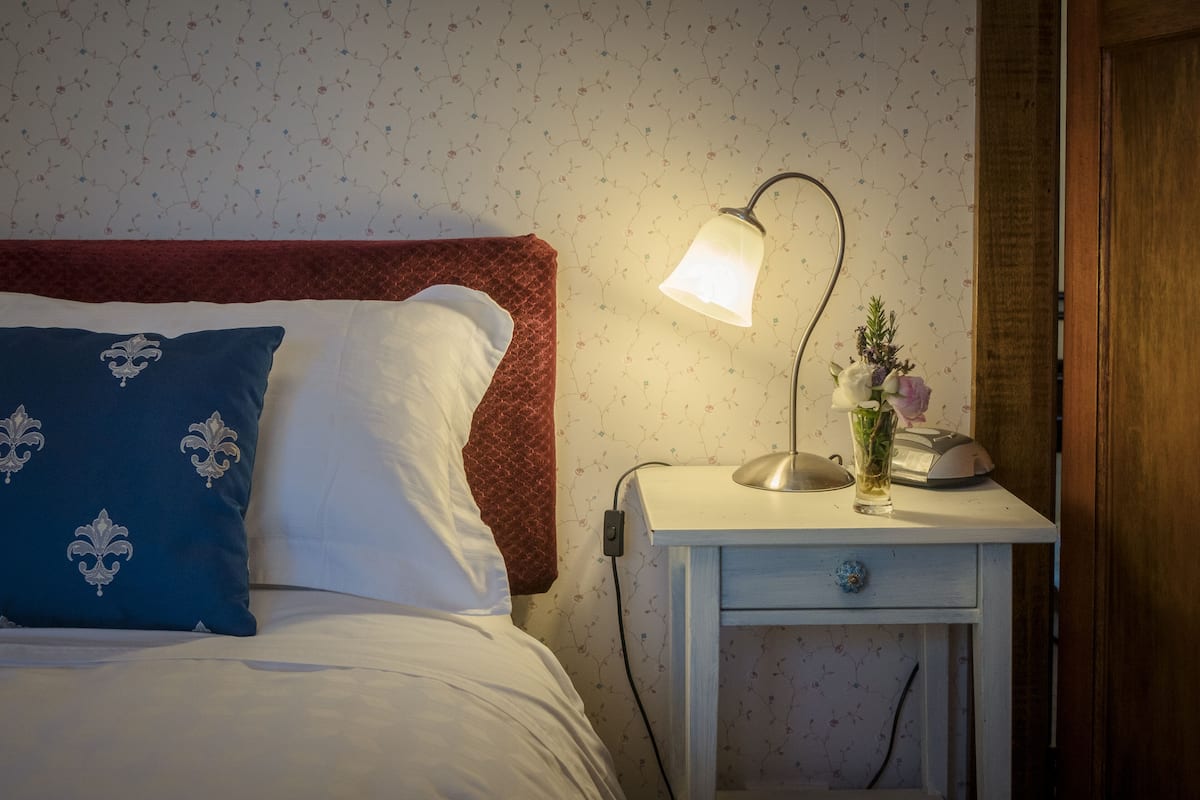 A neatly made bed with a red headboard, a white pillow, and a blue pillow with a floral pattern. There is a bedside table with a lamp, a vase with flowers, and a telephone.