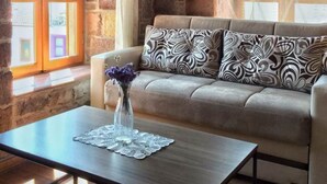 A cozy living room with a stone wall, a sofa with patterned cushions, a coffee table with a vase of flowers, and a window with curtains.