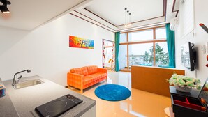 A modern kitchen with a built-in oven, a sink, and a countertop. There is a bright orange sofa and a blue rug in the living area. A painting is on the wall, and a window with curtains is visible. - Sea & Pine Pension (Namhae)