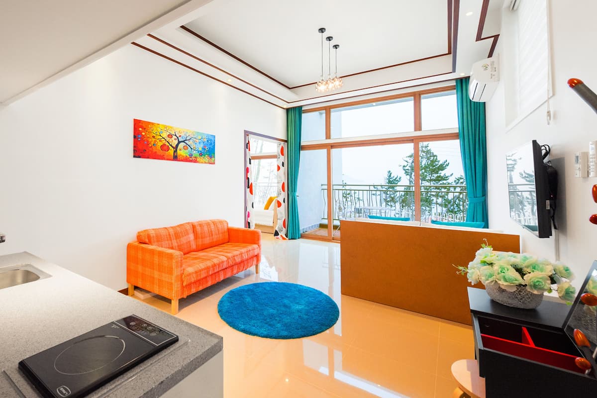A modern kitchen with a built-in oven, a sink, and a countertop. There is a bright orange sofa and a blue rug in the living area. A painting is on the wall, and a window with curtains is visible.
