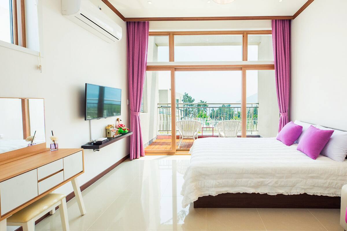 A modern hotel room with a large bed, a TV, a desk, and a balcony with chairs.