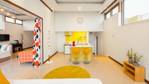 A modern living room with a sofa, a coffee table, and a yellow rug. There is a kitchen area with a yellow countertop and bar stools. - Sea & Pine Pension (Namhae)