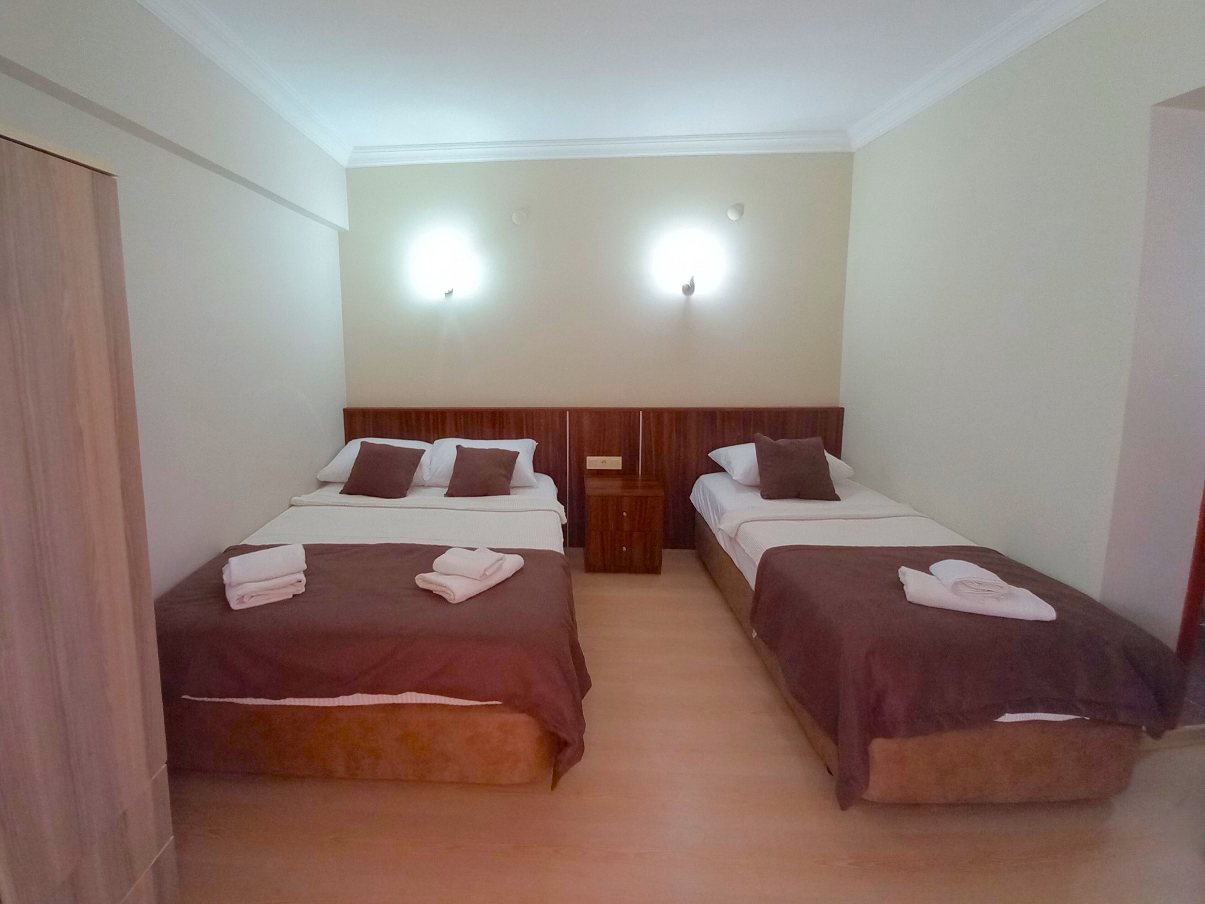 A double room with two beds, each with a wooden headboard, white bedding, and brown throw pillows. There are two wall-mounted lights above the beds.