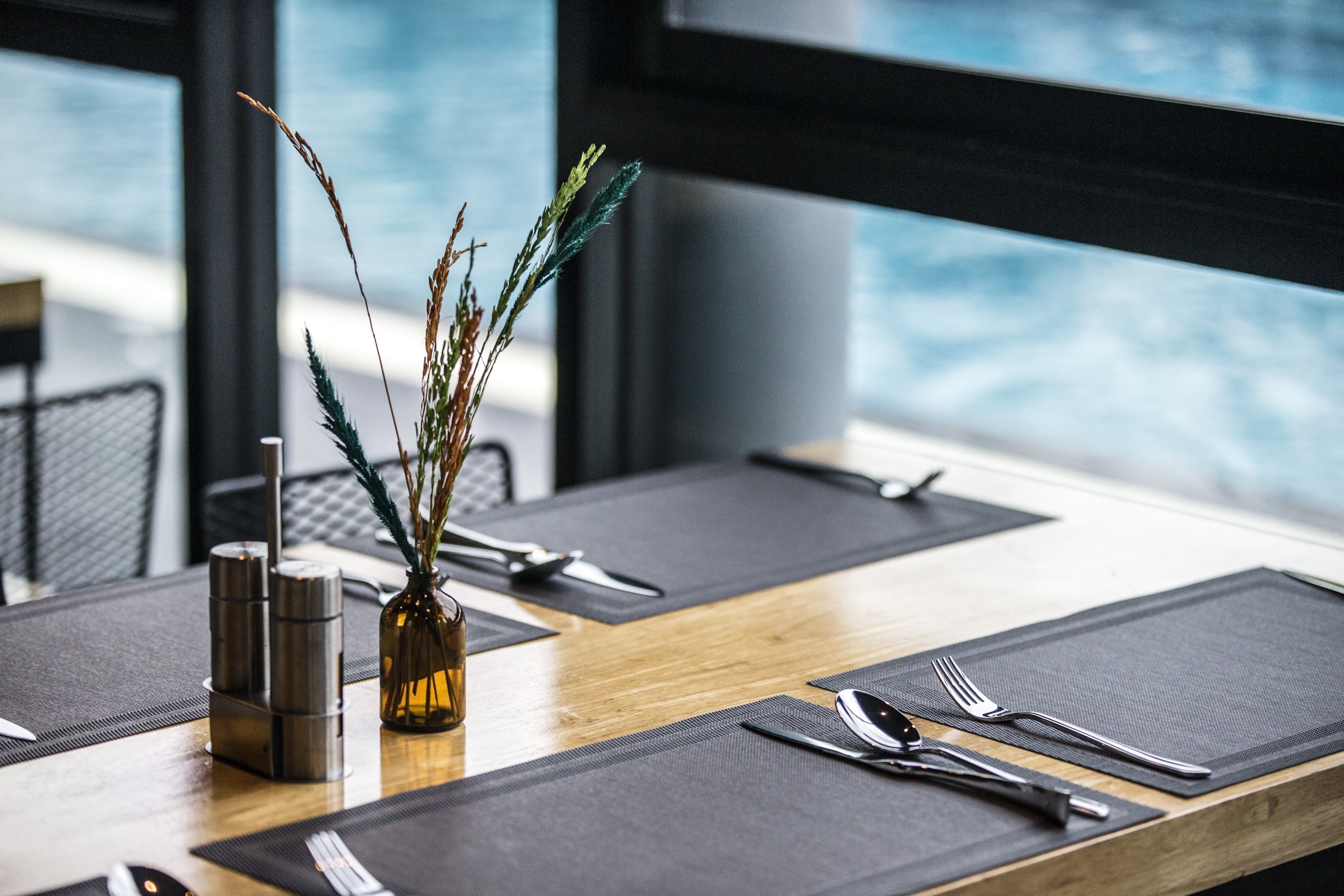 Una mesa de comedor dispuesta con individuales negros, cubiertos y un pequeño jarrón con plantas secas.