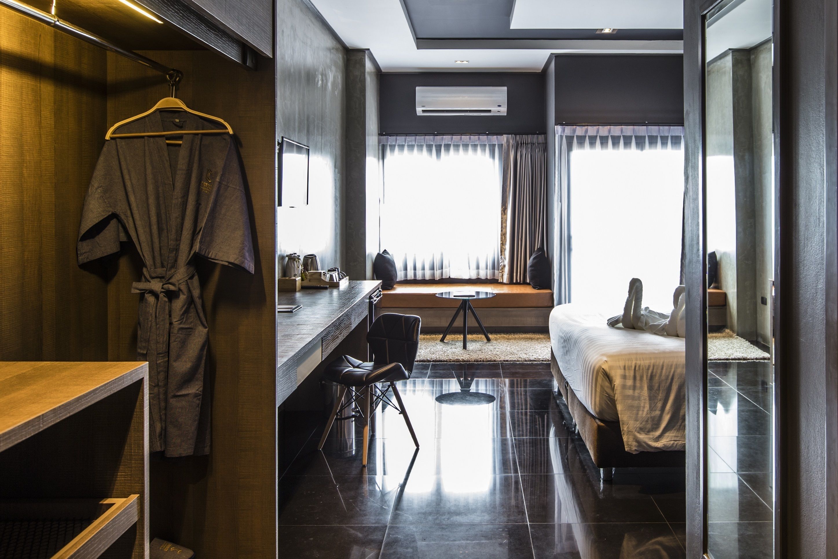 Quarto de hotel moderno com toucador de madeira, roupão pendurado, cama com edredão branco e uma janela grande com cortinas.