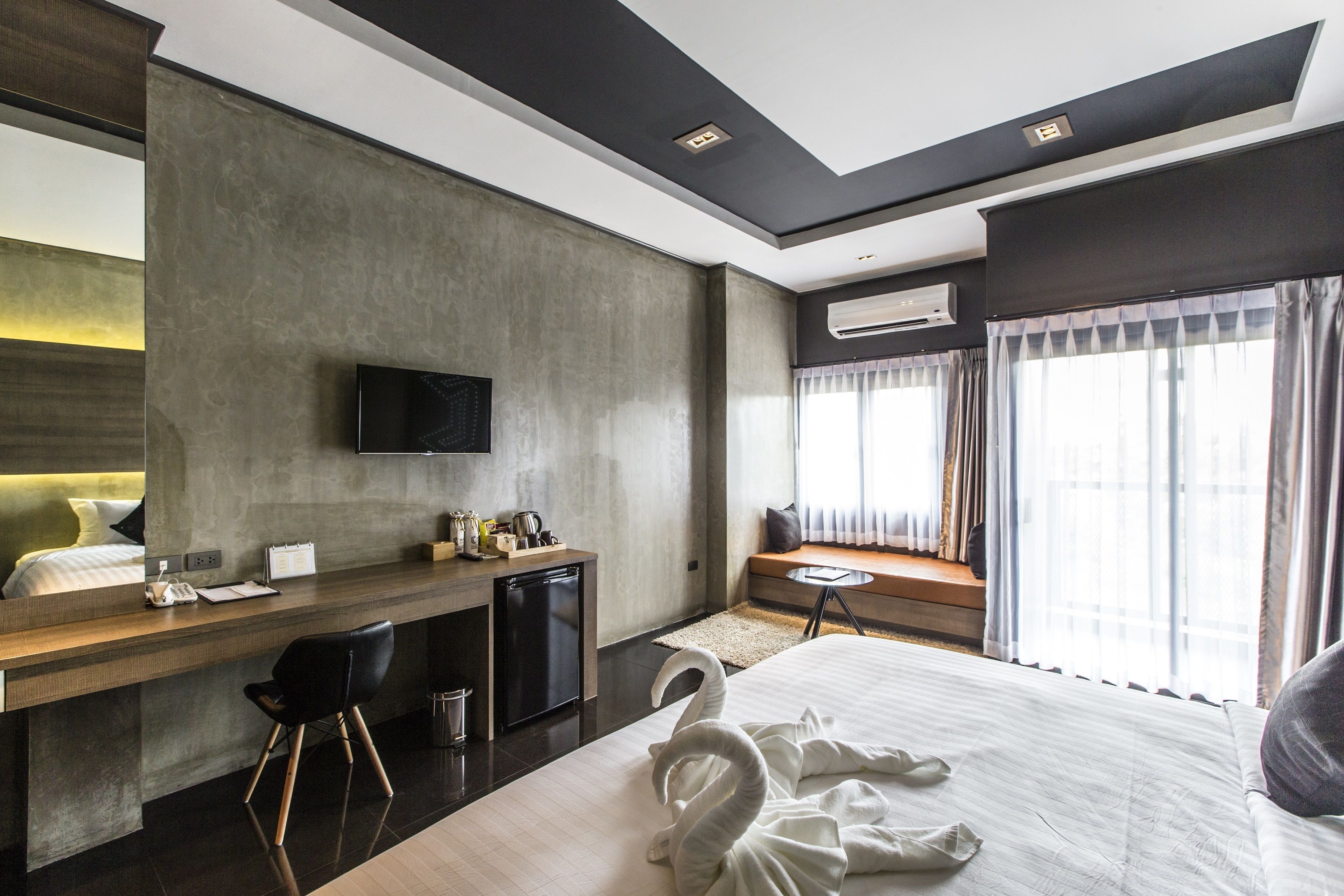 A modern hotel room with a large bed, a desk with a computer, a TV mounted on the wall, and a window with curtains.