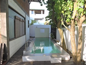 Una pequeña piscina exterior con una escalera de metal, rodeada por un muro blanco y un árbol.
