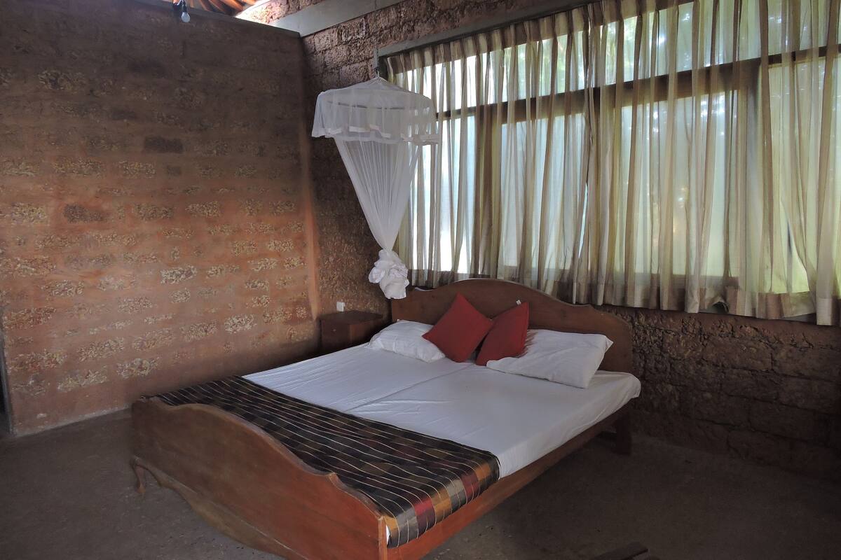 Una cama individual con estructura de madera y cabecera metálica, una colcha blanca y dos almohadas rojas. Hay una mosquitera sobre la cama, y la habitación tiene paredes de ladrillo y una ventana con cortinas de encaje.