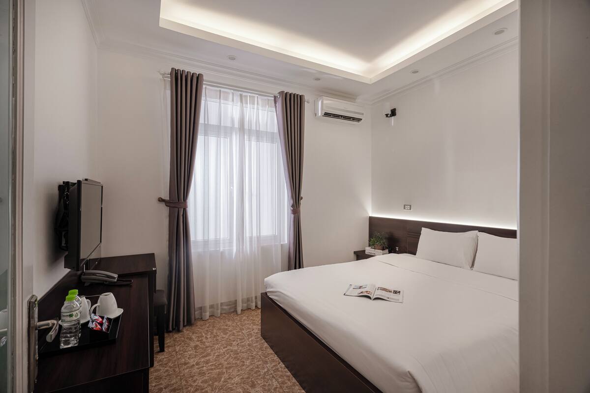 A hotel room with a bed, a bedside table with a lamp, a TV, and a window with curtains.