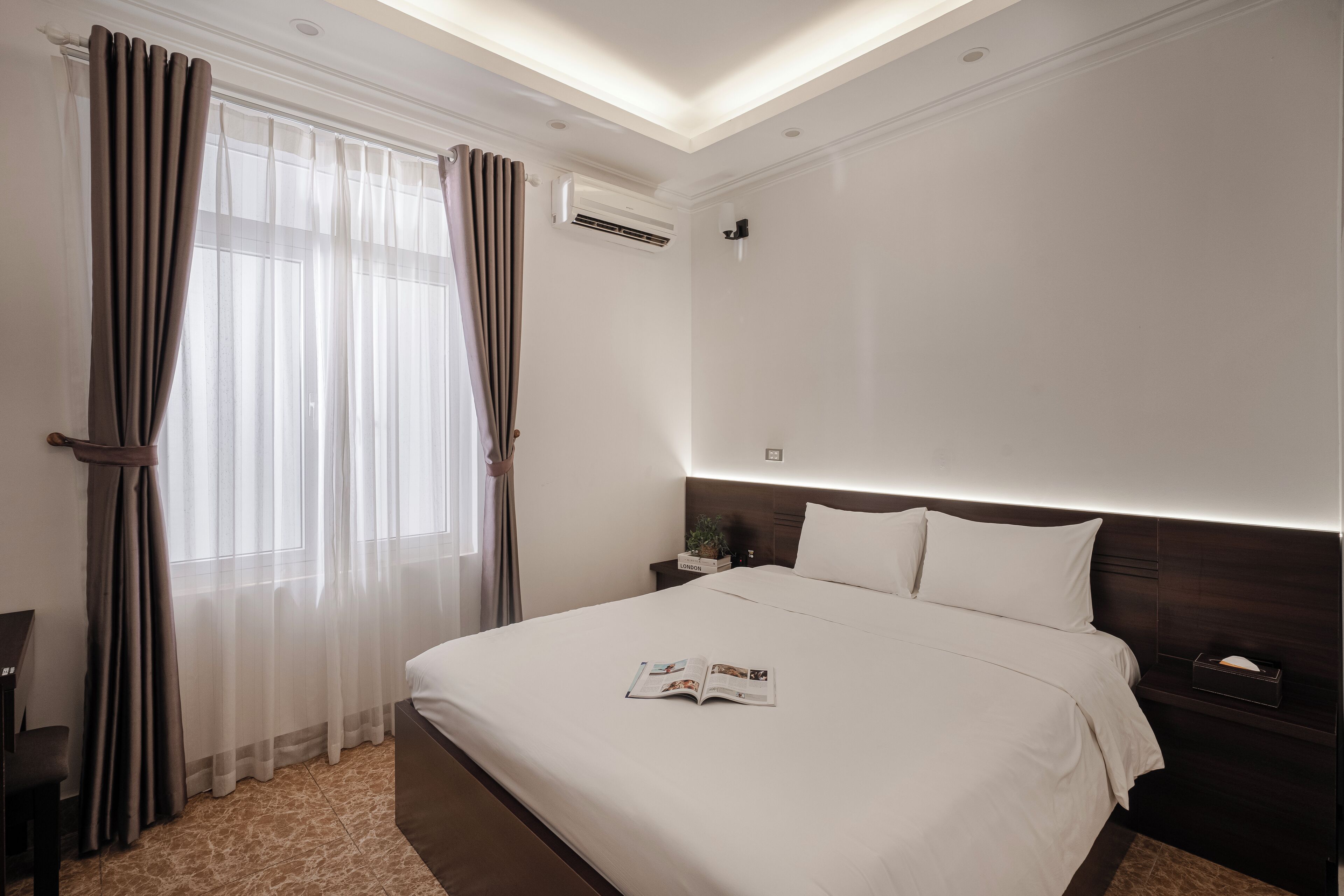 A hotel room with a large bed, a bedside table, a nightstand, a window with curtains, and an air conditioning unit.