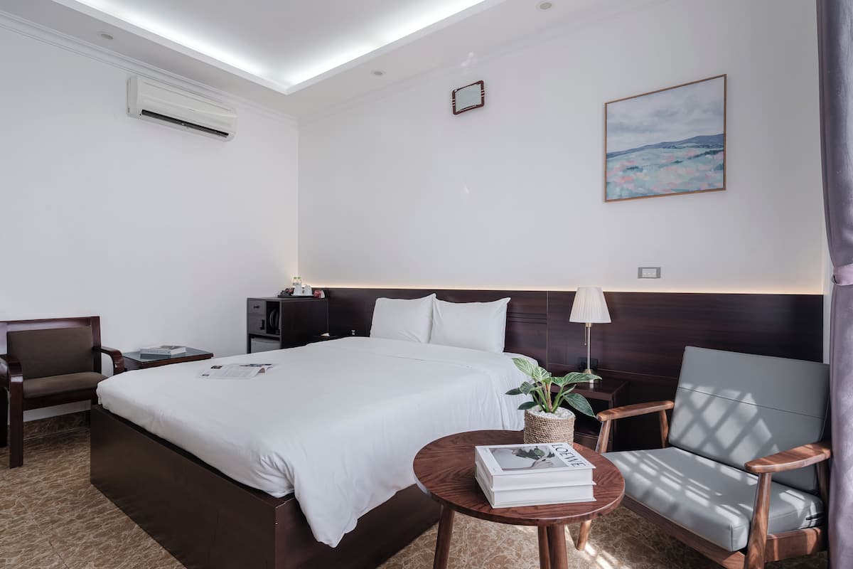 A hotel room with a large bed, a desk with a lamp, a chair, a small table with a book, and a plant.