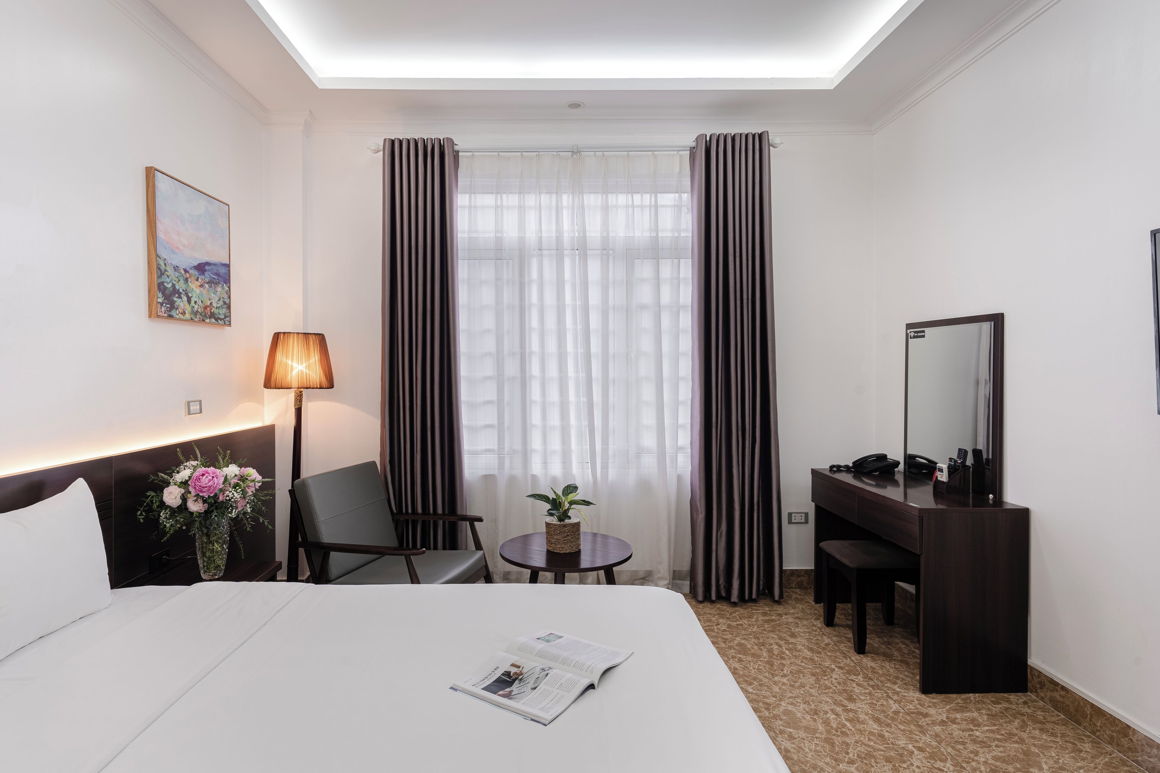 A hotel room with a bed, a desk, a chair, a lamp, a vase with flowers, a mirror, and a window with curtains.