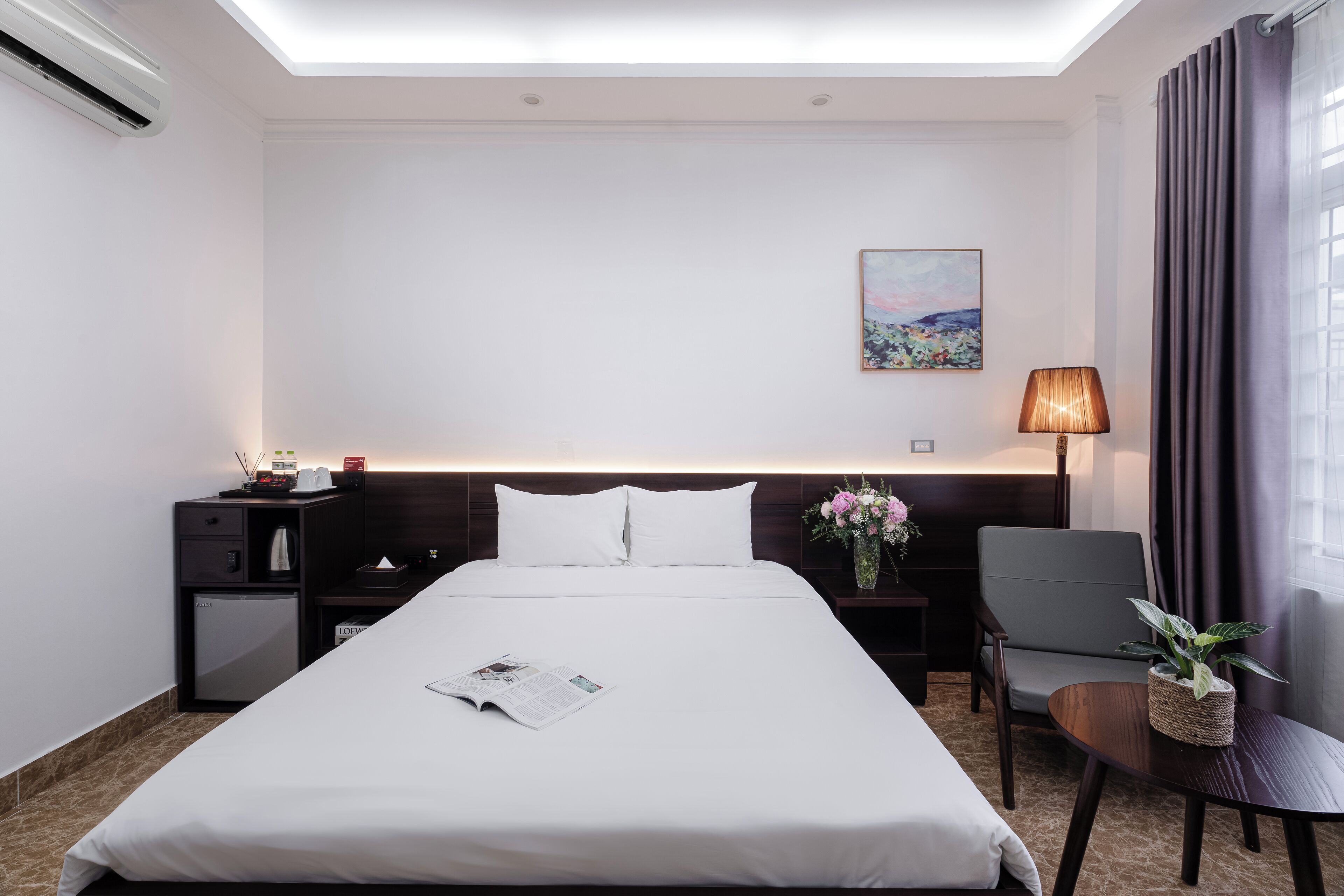 Een hotelkamer met een opgemaakt bed, een klein bureau met een lamp, een stoel en een nachtkastje met een bloemenvaas.