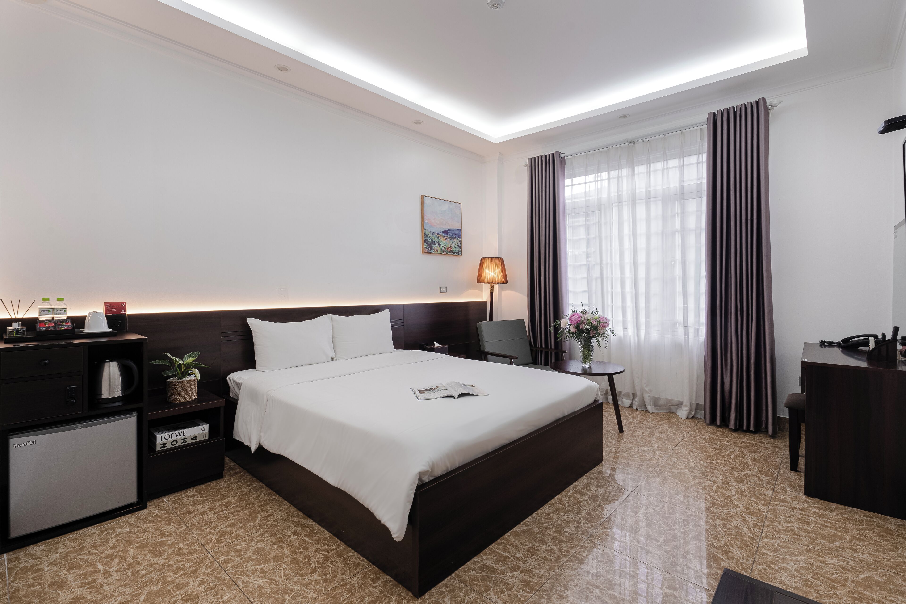A modern hotel room with a large bed, a desk, a small fridge, and a bedside table with a lamp.