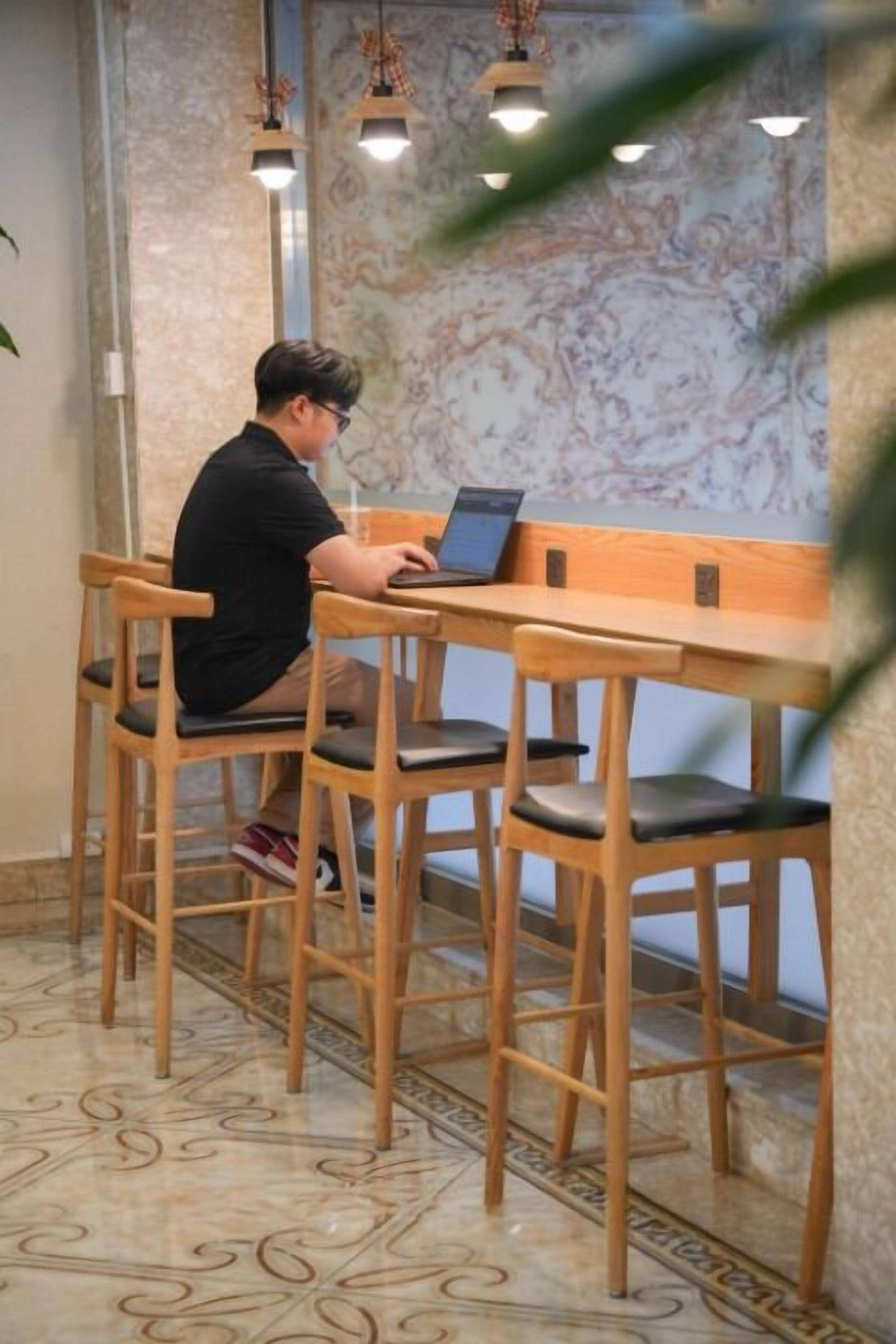 A person seated at a wooden table with a laptop, surrounded by wooden chairs with cushioned seats.